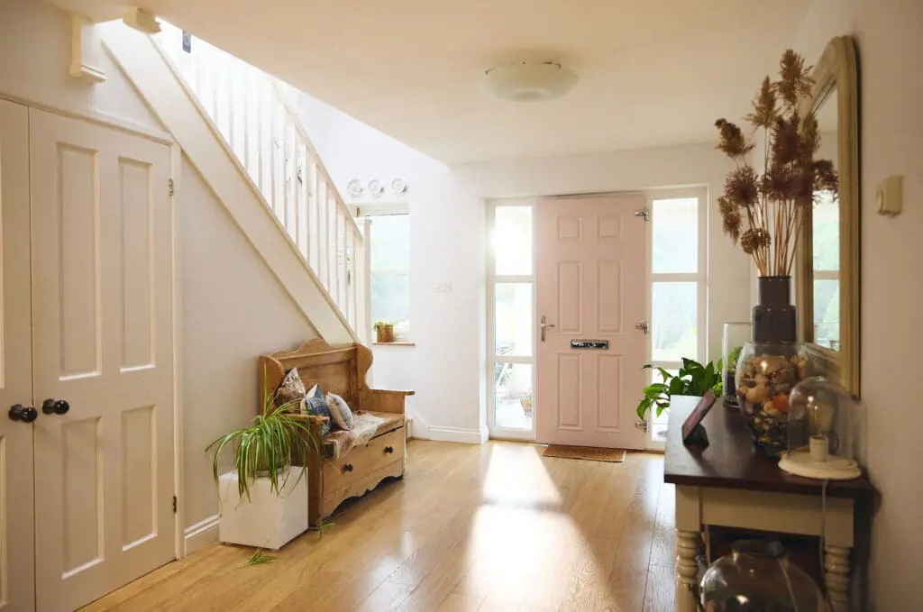 A bright and airy hallway featuring a light pink front door, wooden bench with potted plants, and wooden floor. Natural light filters in through adjacent windows, illuminating the space. A staircase with white railings ascends on the left.