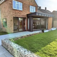 A modern two-story brick house with large sliding glass doors opens to a well-manicured backyard. By the doors, there's a covered patio with seating inside. The garden features a narrow flowerbed bordered by wire mesh stone baskets, and a lush lawn extends to the foreground.