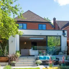 Modern two-story house with large glass doors opening onto a patio. The patio has outdoor seating, potted plants, and steps leading to a green garden, with part of a tree visible on the left. The sky is clear and sunny.