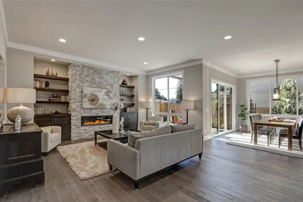 Modern open-concept living and dining area with gray sofas, a stone fireplace, built-in shelves, large windows, sliding glass doors, and light wood flooring; dining table set for four near patio.