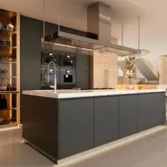 Modern kitchen with a large island, sleek dark cabinets, built-in ovens, open shelving with glassware, under-cabinet lighting, and bar stools. The space is bright with natural light from large windows.