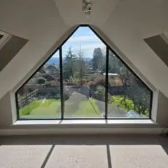 A triangular floor-to-ceiling window offers a clear view of a sunny yard with trees, a driveway, and distant hills. Sunlight streams into the beige-carpeted room, casting sharp shadows on the floor.