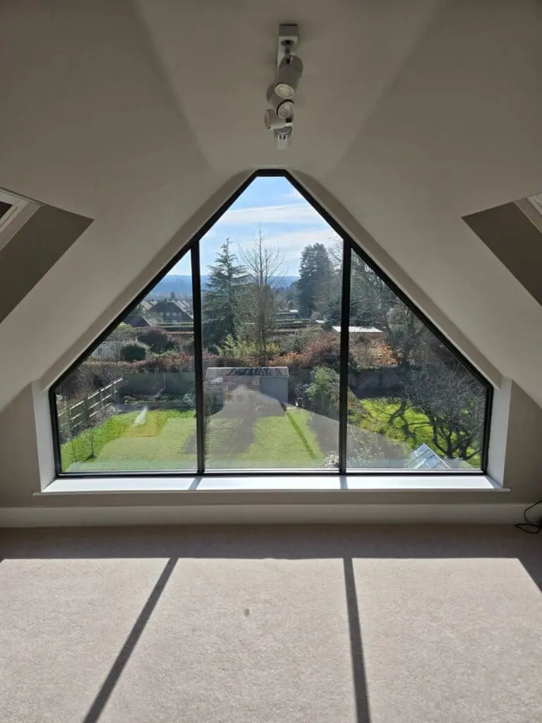 A triangular floor-to-ceiling window offers a clear view of a sunny yard with trees, a driveway, and distant hills. Sunlight streams into the beige-carpeted room, casting sharp shadows on the floor.