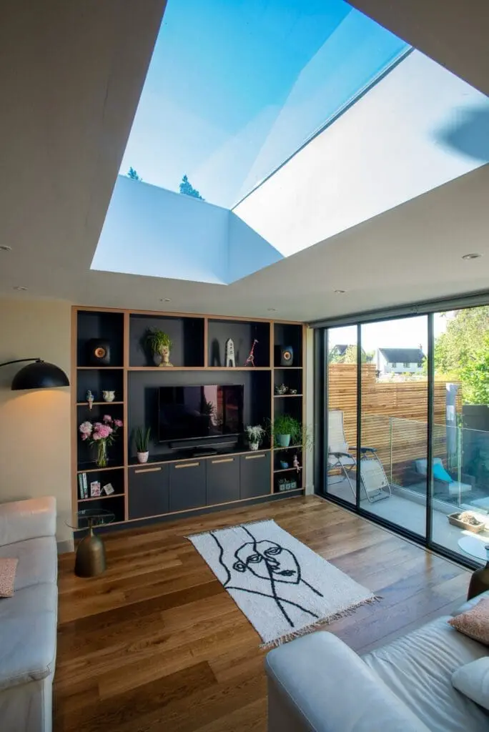 A modern living room with wooden floors, a large skylight, built-in shelves around a TV, white sofas, a line-art rug, and glass doors leading to a patio with greenery outside.