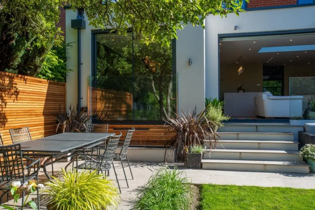 Modern backyard with a patio dining set, plants, and steps leading up to a contemporary house with large glass doors and windows that open to a bright living room with white furniture.