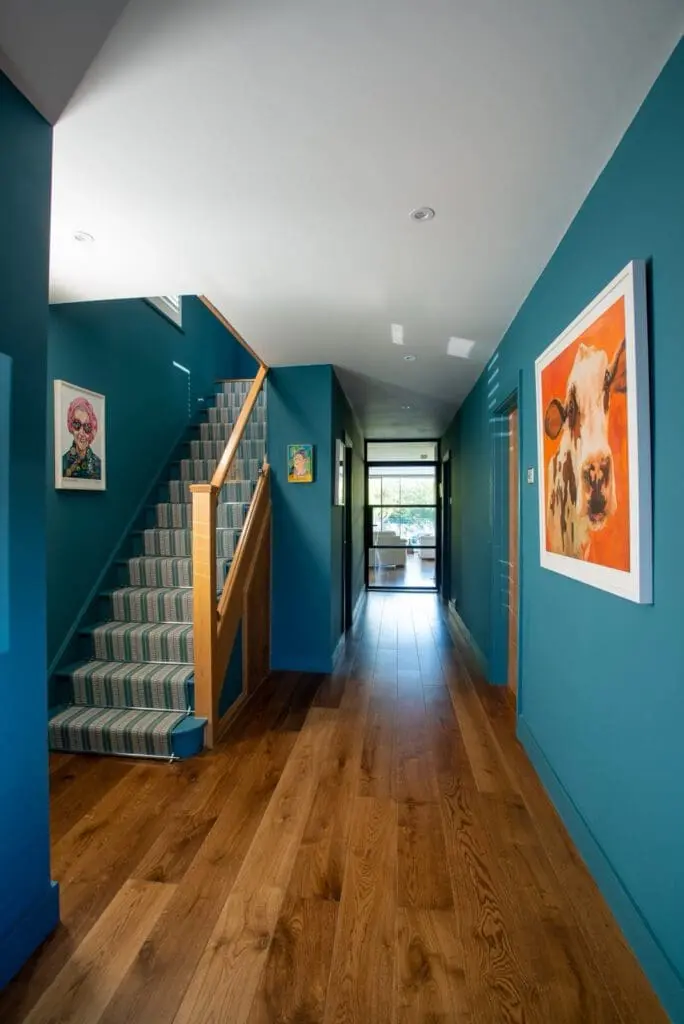 Bright hallway with teal walls, wood floor, and a staircase with striped carpet runner. Colorful artwork, including a cow portrait, decorates the walls. Natural light enters from glass doors at the end.