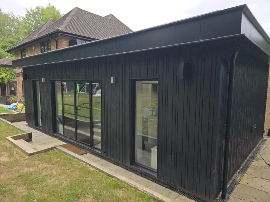 A modern black outbuilding with large glass doors and vertical siding sits on a stone patio in a grassy backyard, near a brick house.