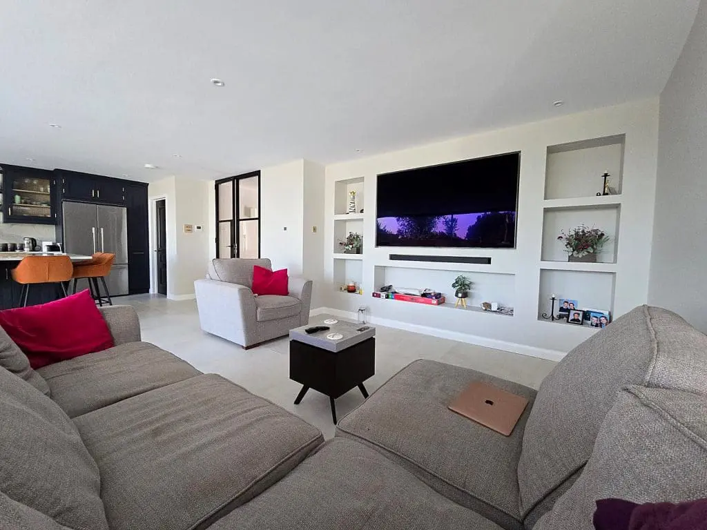 Modern living room with a large gray sectional sofa, armchair, wall-mounted TV, and built-in shelves. A kitchen with black cabinets and bar stools is visible in the background. Pink cushions add a pop of color.