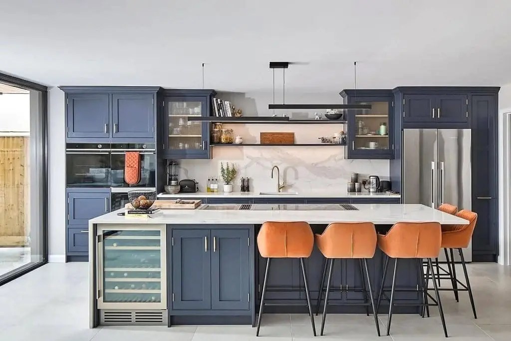 Modern kitchen with blue cabinets, a marble-topped island, built-in appliances, and four orange bar stools. Shelves hold dishes and decor, and large windows let in natural light. Stainless steel refrigerator on the right.