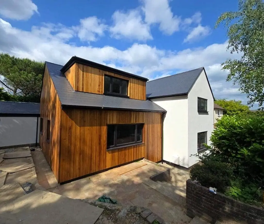 A modern house with a white-painted section and a wooden-clad extension, featuring large windows and a dark slate roof under a partly cloudy sky. The yard is unfinished, with bare ground and some greenery nearby.