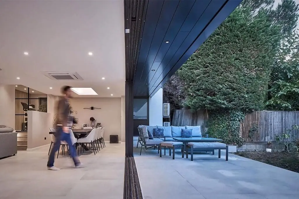 A modern open-plan living space seamlessly extends to an outdoor patio with sofas and trees. A person is walking inside near a long dining table, with large glass doors fully opened to the garden.