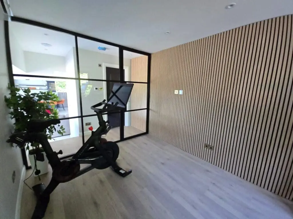 A modern home gym room with light wood flooring and vertical slat wall panels, featuring an exercise bike, indoor plant, and glass partition separating the space from the adjacent area.