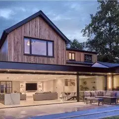 Modern two-story house with wood and brick exterior, large windows, and fully open sliding glass doors revealing a spacious, well-lit living area and outdoor patio with seating at dusk.