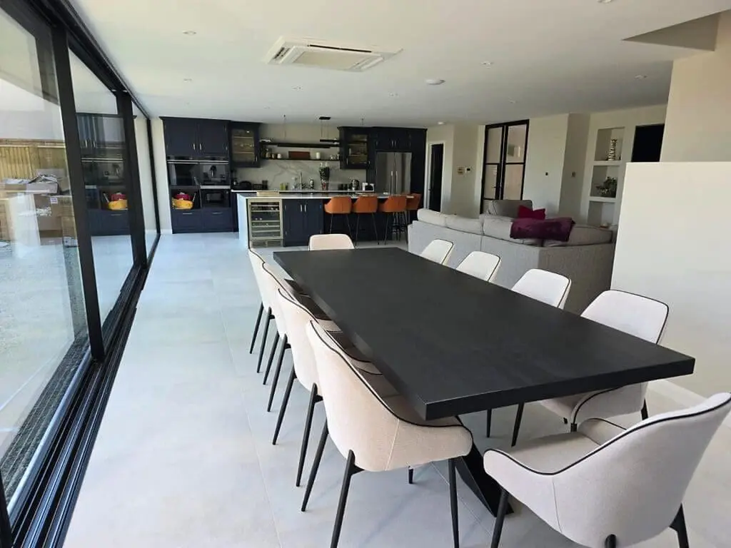 Modern open-plan kitchen and dining area with a long black table surrounded by light upholstered chairs, dark kitchen cabinets, orange bar stools, and a gray sofa in the background. Large windows line one side.
