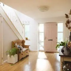 A bright hallway with wooden floors, a pink front door, a wooden bench with cushions and plants, a staircase, and a console table decorated with glass jars and dried flowers. Natural light streams in from the window and door.