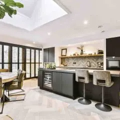 Modern kitchen with black cabinets, a large island with bar stools, built-in ovens, and open shelving. Adjacent dining area with round table and chairs, large windows with shutters, and a skylight above.