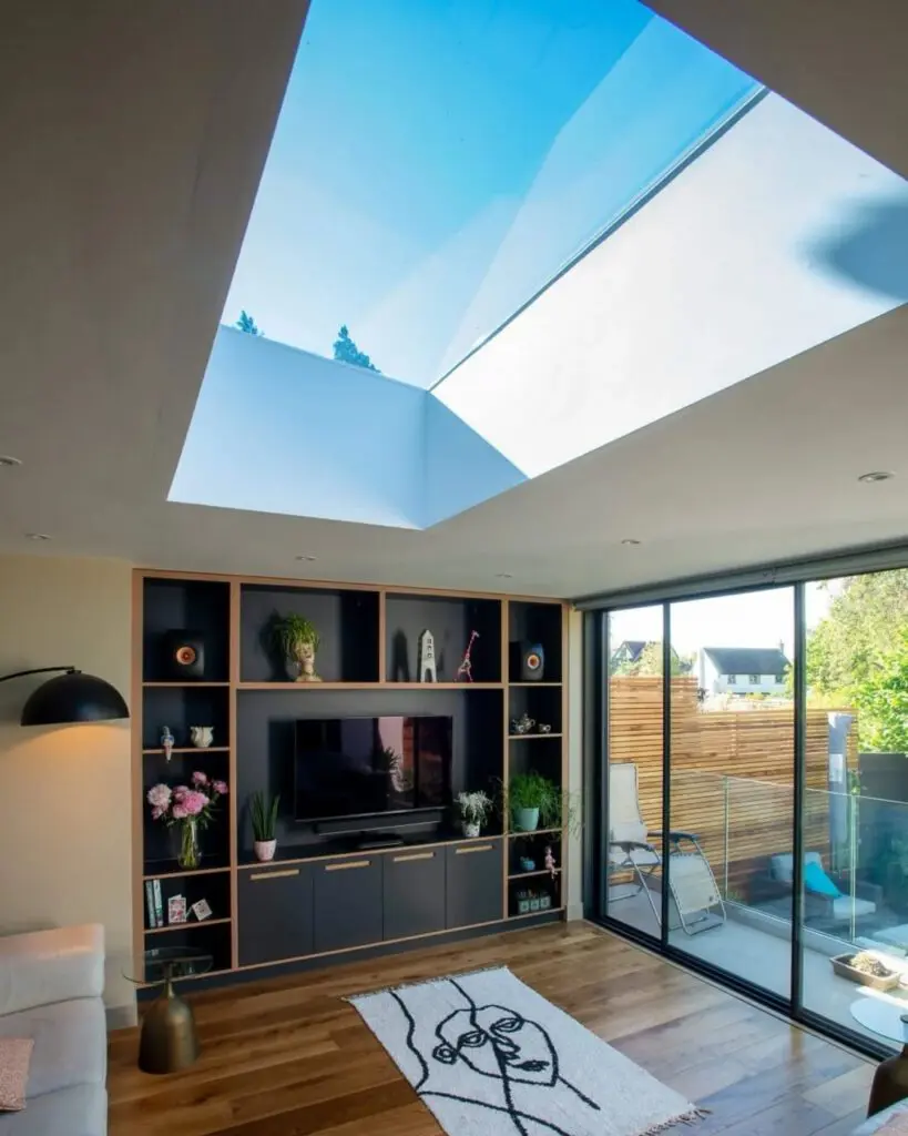 Modern living room with a skylight, built-in shelving around a TV, sliding glass doors leading to a balcony, wooden floors, and a white rug with a black abstract face design.