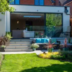 A modern house with large glass doors opens to a patio featuring outdoor seating, potted plants, steps, and a well-kept lawn with lush greenery under a sunny sky.