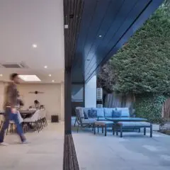 A person walks through a modern open-plan home with large sliding doors, revealing a patio with outdoor seating and lush trees in the backyard.