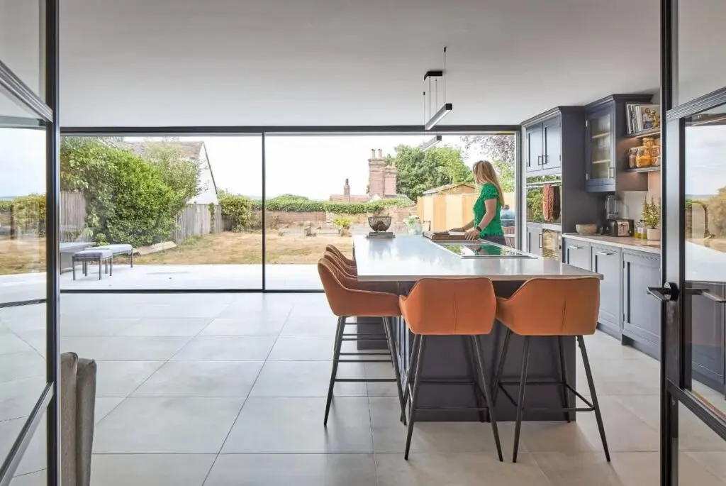 Modern kitchen with large glass windows, an island with brown barstools, and a woman in a green dress standing by the counter. The view outside shows a yard and distant buildings.
