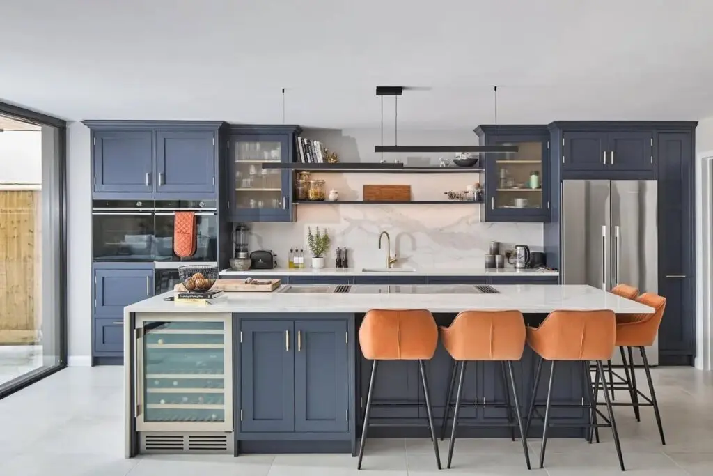 Modern kitchen with blue cabinets, a large island with a white countertop, and four orange barstools. Stainless steel appliances, open shelves with decor, and a built-in wine cooler are visible. Natural light fills the space.
