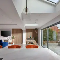 Modern open-plan living space with glass pendant lights, orange chairs, a marble countertop, a blue sofa, wall-mounted TV, dining table, large sliding glass doors, and a stone patio outside. Natural light fills the room.