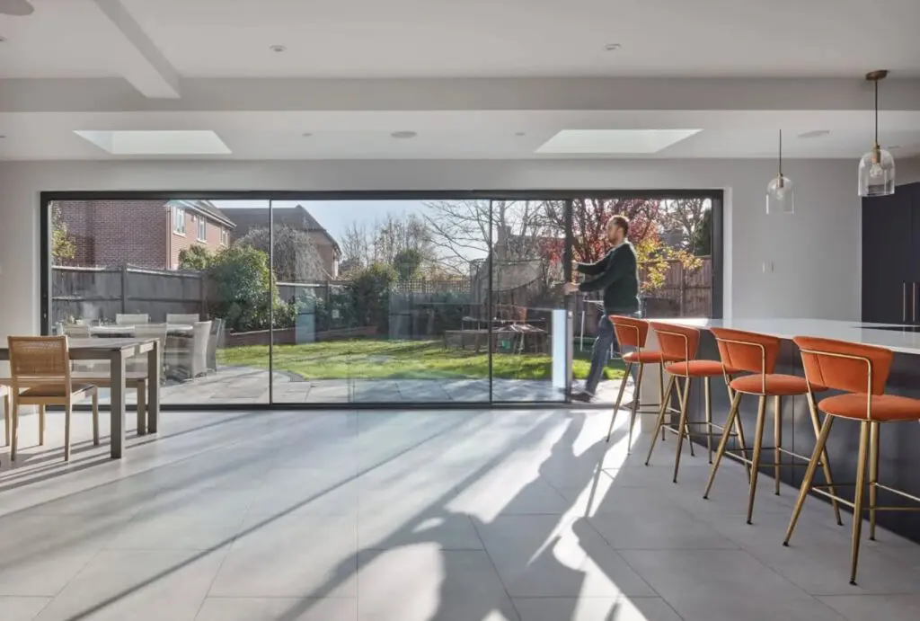 A modern kitchen with orange barstools and large sliding glass doors opening to a sunny backyard, where a person is standing outside near the grass.