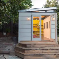 A small modern shed with white paneling, glass double doors, and wooden steps sits in a garden surrounded by trees and plants. Warm yellow light glows from inside, showing shelves and a chair.