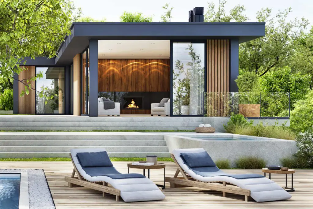 Modern house with large windows, wood accents, and a lit fireplace. Two cushioned lounge chairs and small tables sit on a poolside deck, surrounded by greenery and trees.