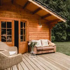 Wooden cabin with glass doors, a wicker chair, a brown couch with a blanket, flowers on a small table, and a green lawn with trees in the background.