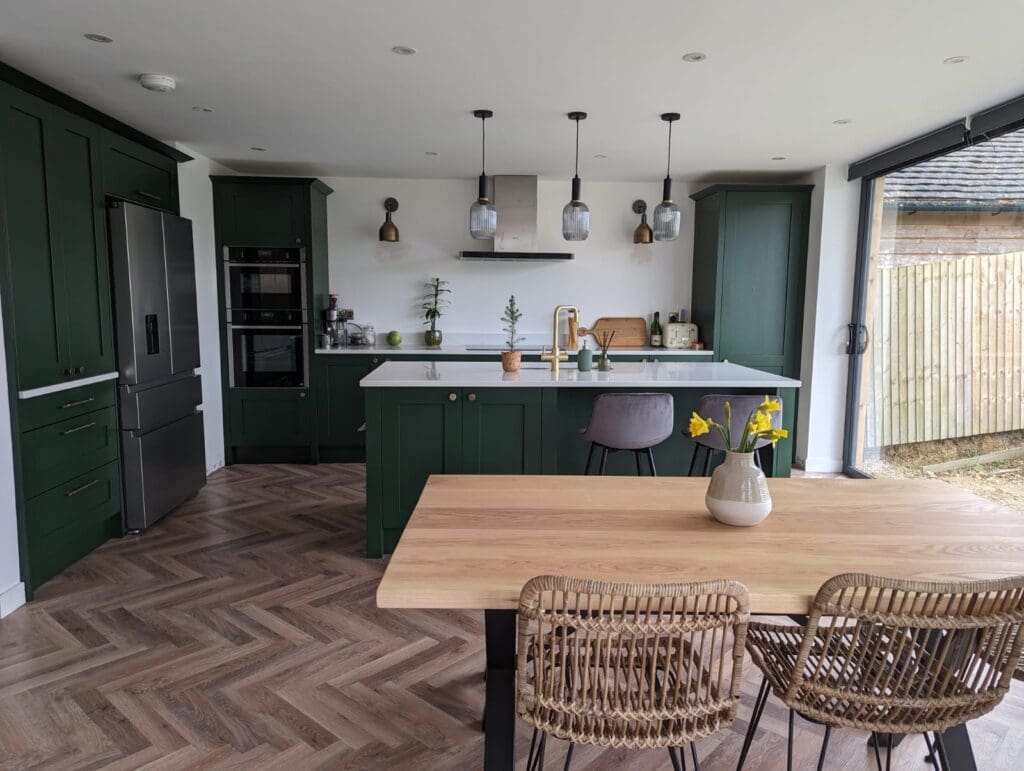 Modern kitchen with green cabinetry, built-in double oven, and stainless-steel fridge. Central island with plants, a wooden countertop, and a gray chair. Three pendant lights hang above. Dining table with woven chairs, a vase of yellow flowers, and large windows.
