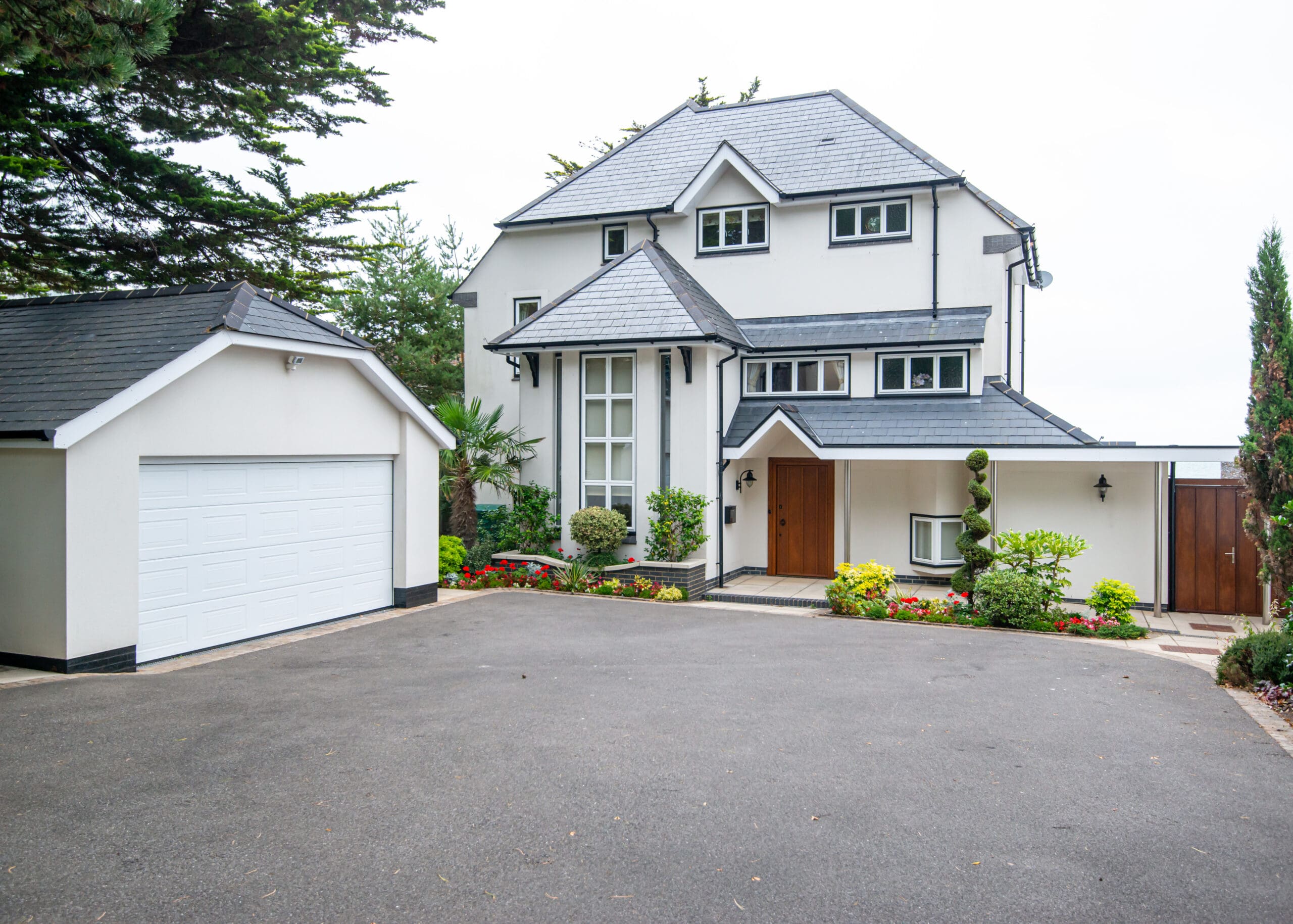 A large, modern white house with grey roof tiles surrounded by greenery. The house has a main entrance with a wooden door, multiple windows, and an attached garage on the left. A well-maintained driveway leads to the entrance, with a garden featuring various plants and shrubs.