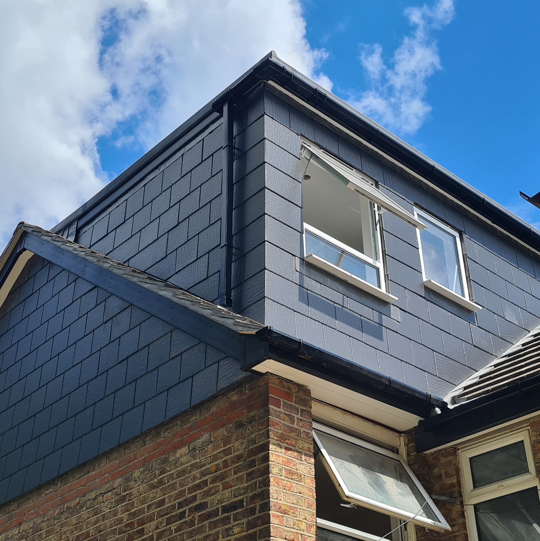 A building exterior with a recently constructed dark-shingled dormer extension. The dormer features two open windows. The sky above is partly cloudy with patches of blue. A section of building scaffolding is visible on the right side.