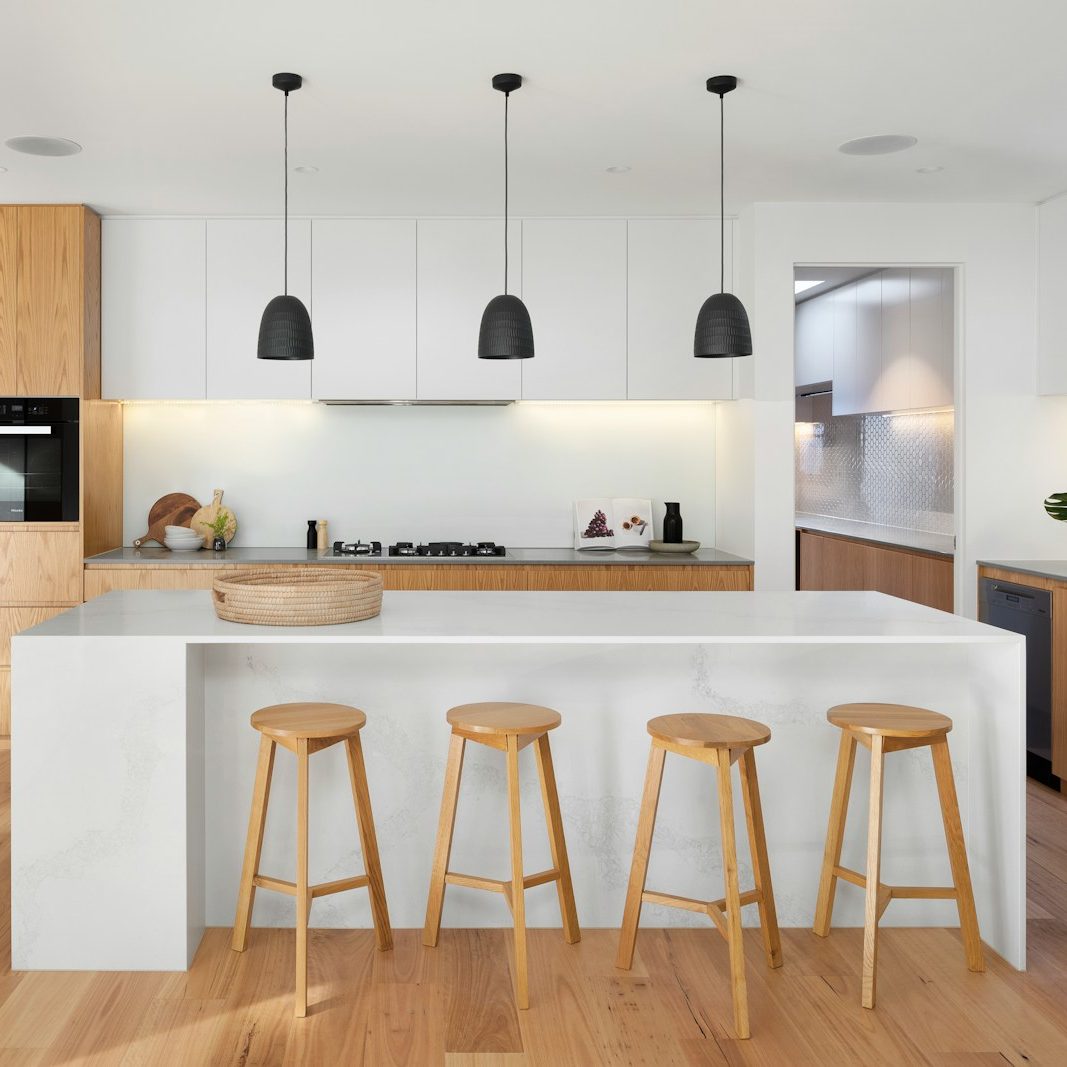 Modern kitchen with light wood flooring, white countertops, and cabinets. A large island with three wooden stools faces the open space. Pendant lights hang above the island. Stainless steel appliances, green plant, and decor items enhance the minimalistic design. Stairs leading upstairs are visible in the background.