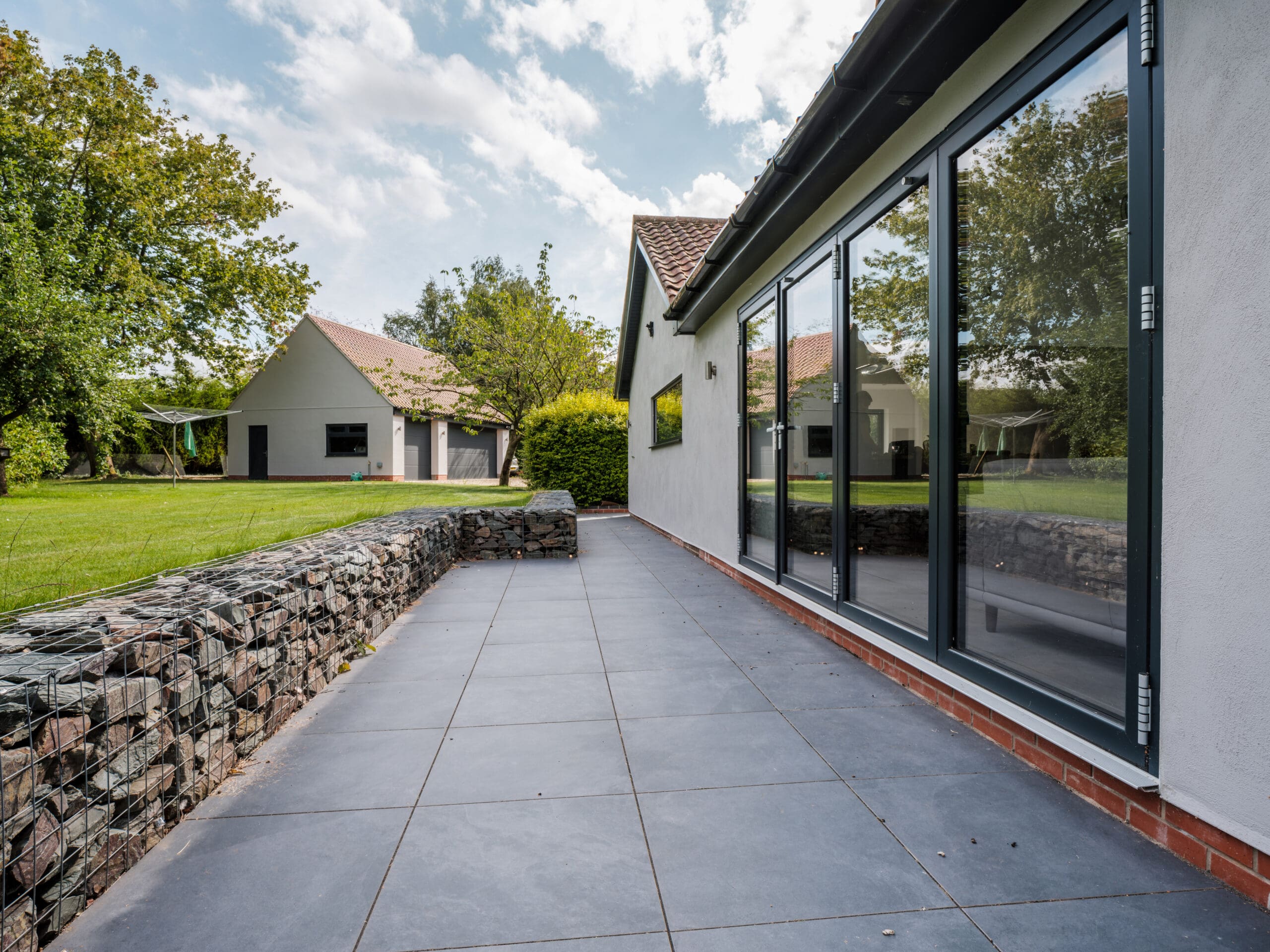 Modern patio with large gray tiles and glass sliding doors, bordered by a gabion stone wall. In the background, a spacious green lawn and another house surrounded by trees under a partly cloudy sky.