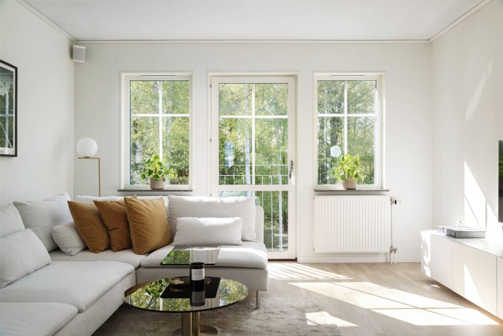 A bright, modern living room with a large white sectional sofa, beige and brown cushions, a round black coffee table, and potted plants on the windowsill. Sunlight streams through three tall windows and a glass door.