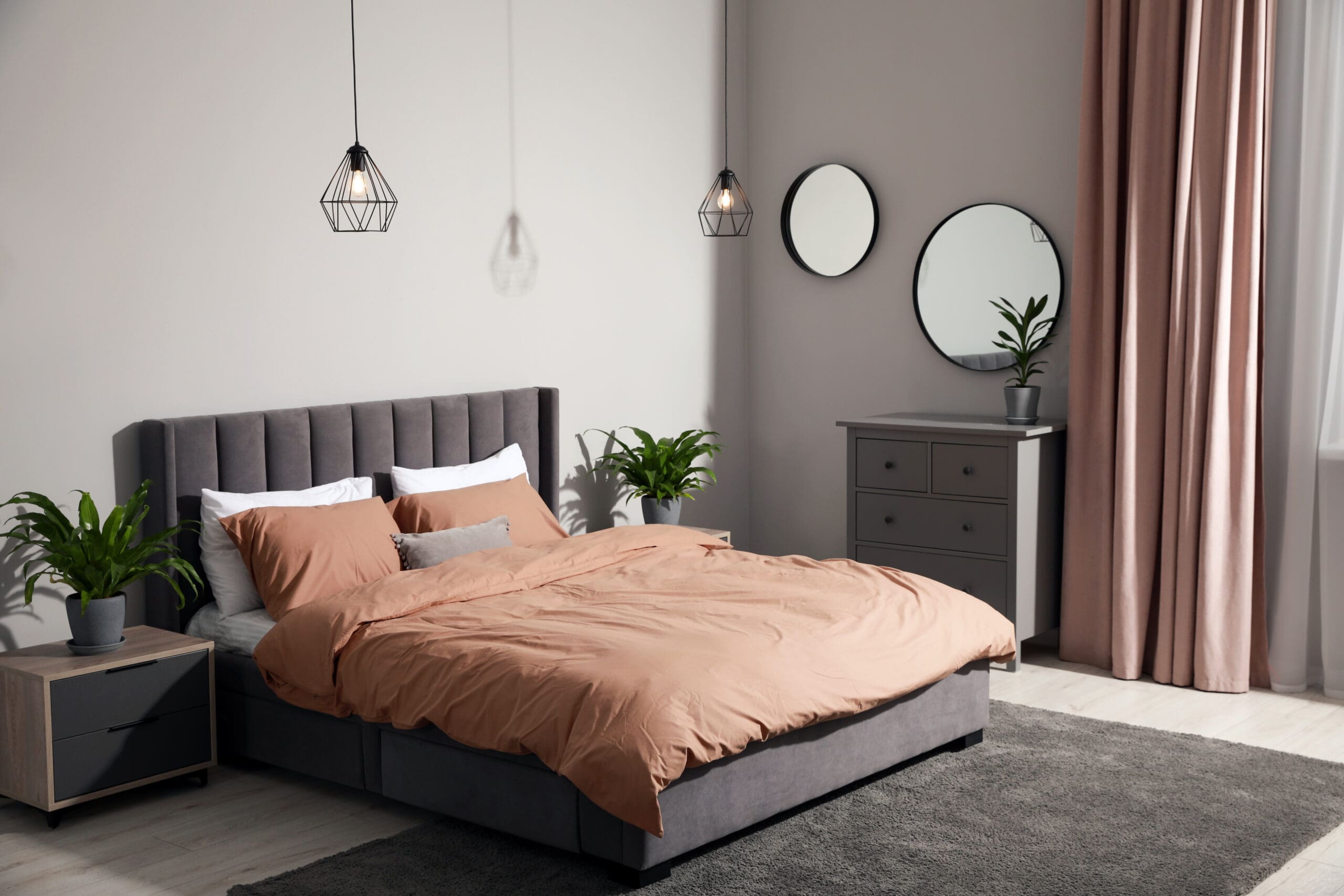 Modern bedroom with a grey upholstered bed and peach bedding, flanked by potted plants on nightstands. Two round mirrors and a dresser are on the back wall, with pink curtains by a window letting in natural light.