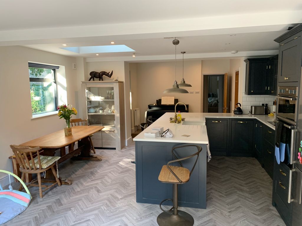 Modern kitchen with dark cabinets, an island with bar stools, and a light wood dining table and chairs. Sunlight streams through a window and skylight. A glass cabinet and piano are in the background. Flowers sit on the table.