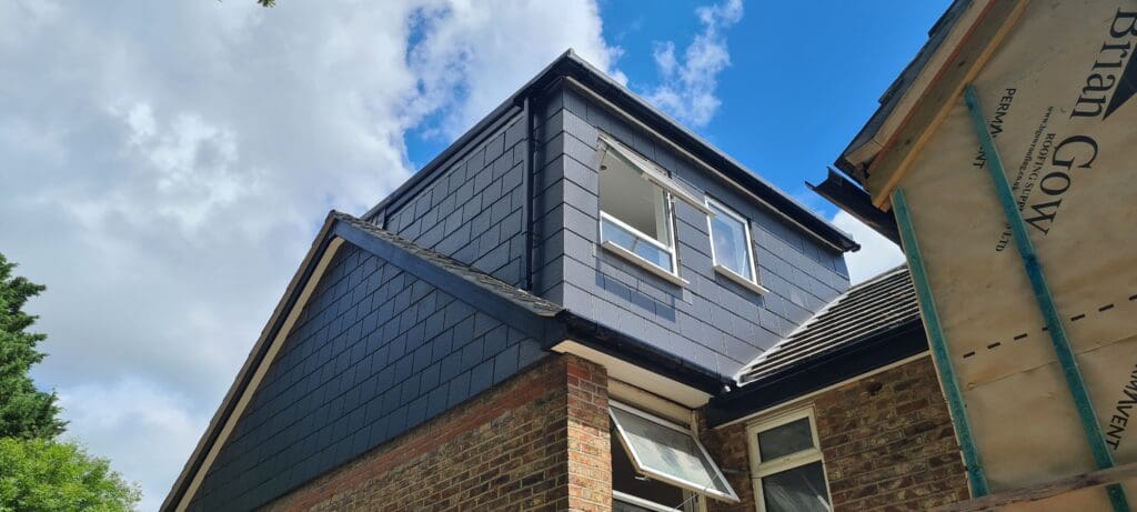 A newly constructed house extension with dark cladding and open windows under a blue sky with some clouds. Part of the original brick structure and roof, along with scaffolding and a construction banner, are visible.