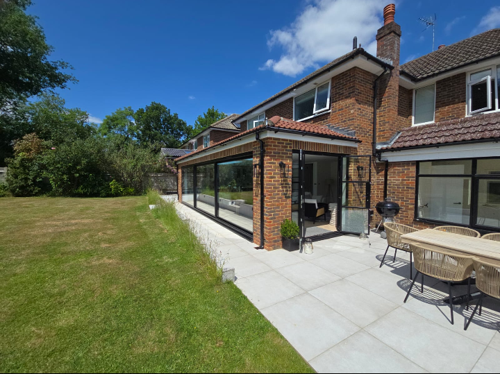 Modern brick house with large glass sliding doors opening to a patio with outdoor seating and a barbecue grill. The patio overlooks a spacious, well-kept lawn with trees in the background under a blue sky.