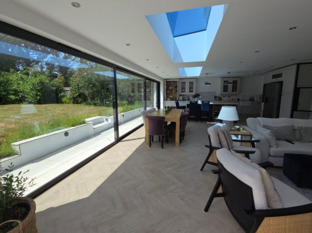 A modern open-plan living and dining area with large glass sliding doors overlooking a green garden, skylights in the ceiling, and contemporary furniture including a wooden dining table and cushioned chairs.