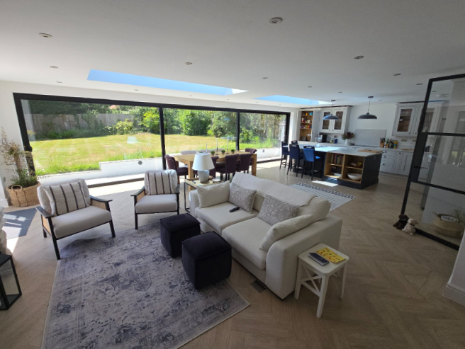 A spacious, modern open-plan living area with a white sofa, armchairs, a rug, and ottomans, adjacent to a dining table and a kitchen. Large glass doors overlook a green garden, letting in natural light.