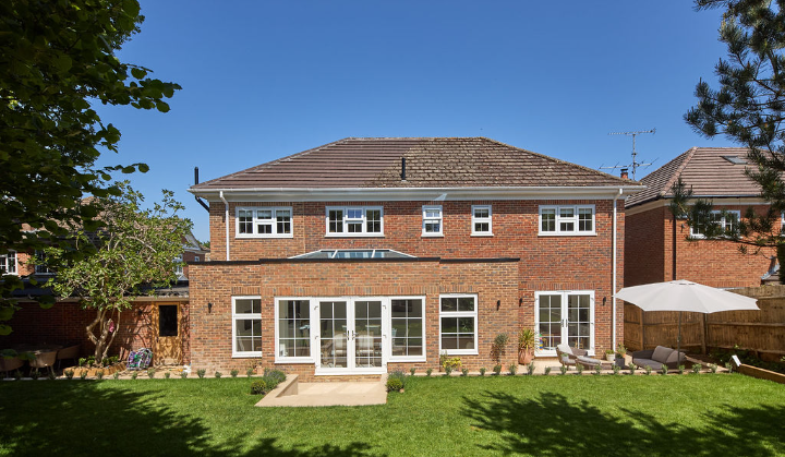A two-story red brick house with white-framed windows and glass doors, facing a sunny, neatly mowed backyard with a patio, small potted plants, and a white umbrella on the right side.