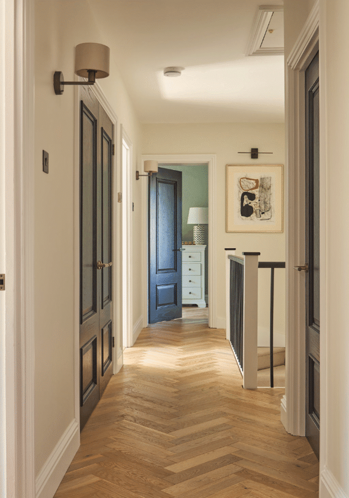 A bright hallway with herringbone wooden floors, cream-colored walls, dark wooden doors, and a black railing. One door is open, revealing a room with a white dresser, lamp, and framed abstract art on the wall.