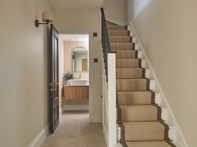 A hallway with a beige-carpeted staircase on the right, cream walls, and a dark door leading to a modern bathroom with a round mirror, sink, and wooden vanity. A wall sconce is mounted above the door.