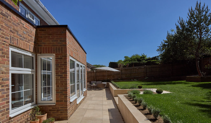 A modern brick house with large windows opens onto a spacious patio. The patio features outdoor seating, a sun umbrella, landscaped grass, small shrubs, and is bordered by a fence and tall trees under a clear blue sky.