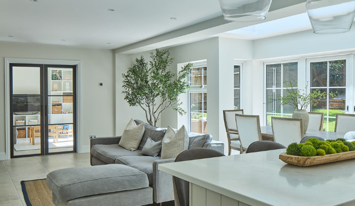 Modern, bright open-plan living and dining area with grey sofas, a large ottoman, dining table with chairs, indoor potted tree, and large windows letting in natural light. A kitchen island is in the foreground.