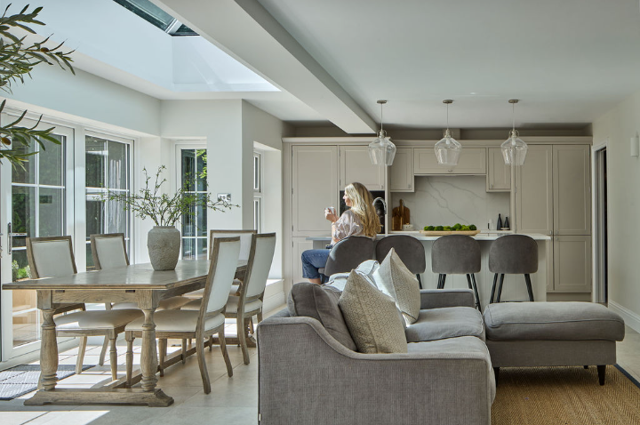 A bright, modern open-plan kitchen and living area with neutral tones. A woman sits at the kitchen island drinking from a mug. Sunlight streams in through large windows near a dining table with chairs.