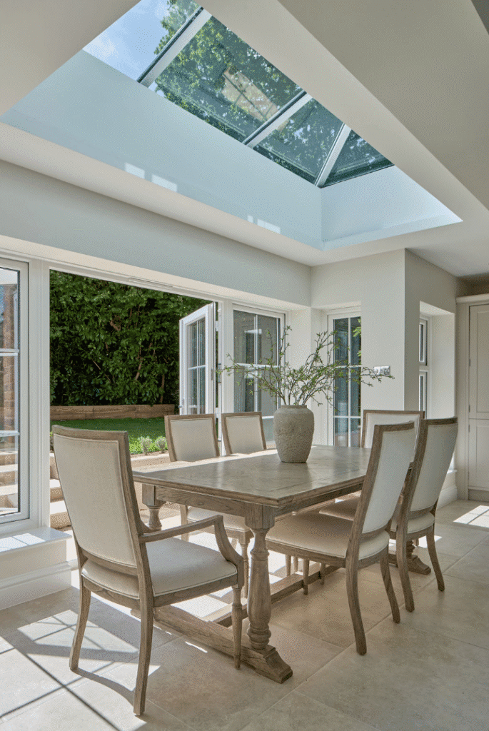 A bright dining area with a wooden table, six upholstered chairs, large windows, and a glass ceiling skylight. Sunlight fills the room, and a vase with branches sits on the table. Greenery is visible outside.