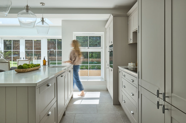 A bright, modern kitchen with white cabinets and countertops, large windows letting in natural light, and a woman walking past the sink area, slightly blurred in motion. A bowl of green apples sits on the island.